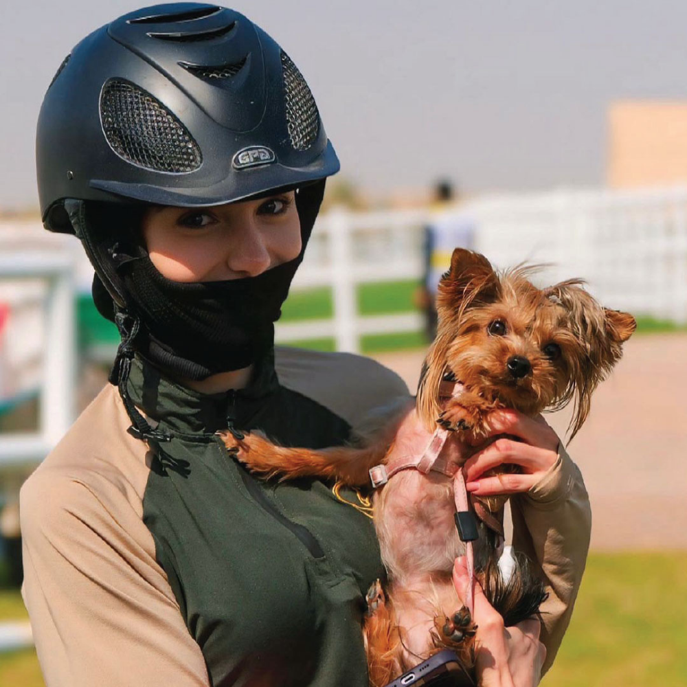 A rider wearing a helmet holds a small dog, showcasing the N3F Riding Mask in a sunny outdoor equestrian setting.